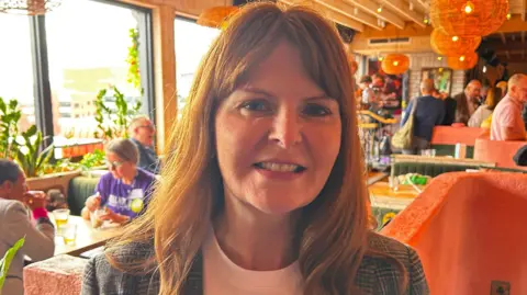 A woman with red hair and wearing a grey checked blazer. She is wearing a white t-shirt. There is a room behind her with people sitting at tables.