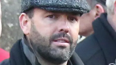 Collins Photo Agency Daniel Kinahan, a man with short dark hair and a stubbly beard, pictured at a funeral in 2016. He is wearing a grey peaked cap, a black shirt and a black coat with the collar pulled up.