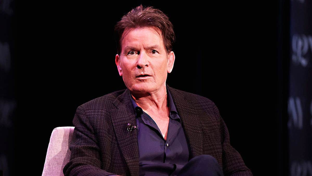 Charlie Sheen in dark clothing looking surprised during an interview.