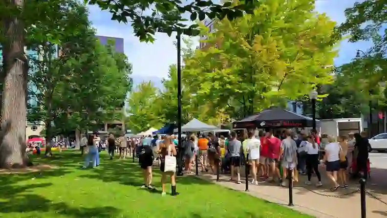 Long line of shoppers at a market tent under leafy trees