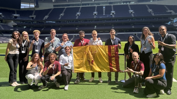Group of ASU students pose on a field in a stadium while holding up a large flag featuring an ASU pitchfork.