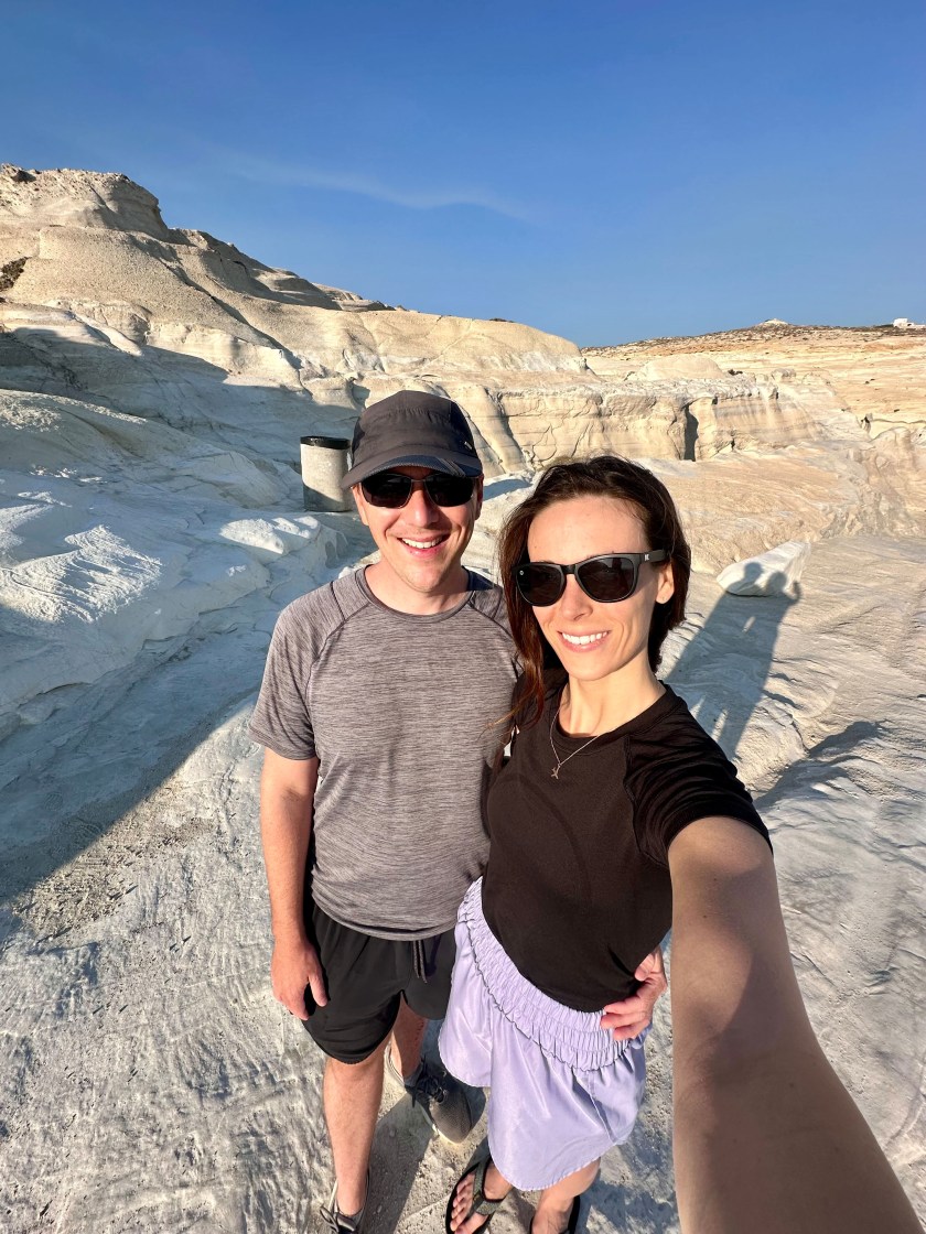 A man and woman pose for a selfie on sunny day in a rocky landscape in Greece.