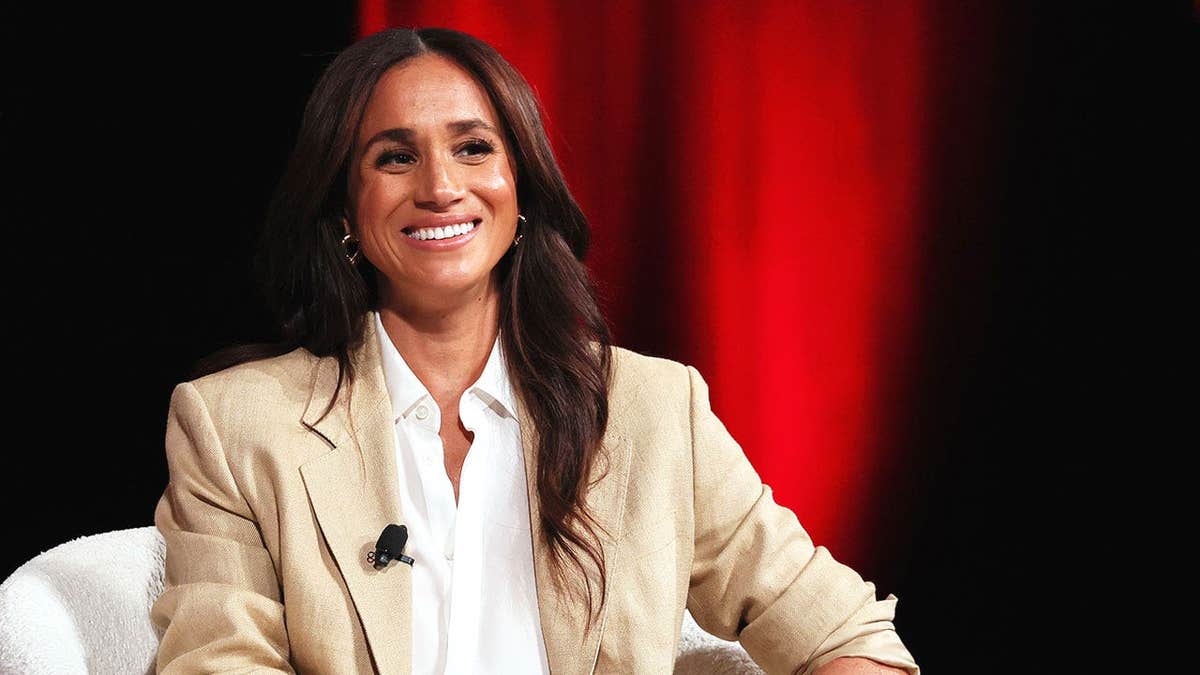 Meghan Markle wearing a beige blazer and smiling in front of a red velvet curtain.