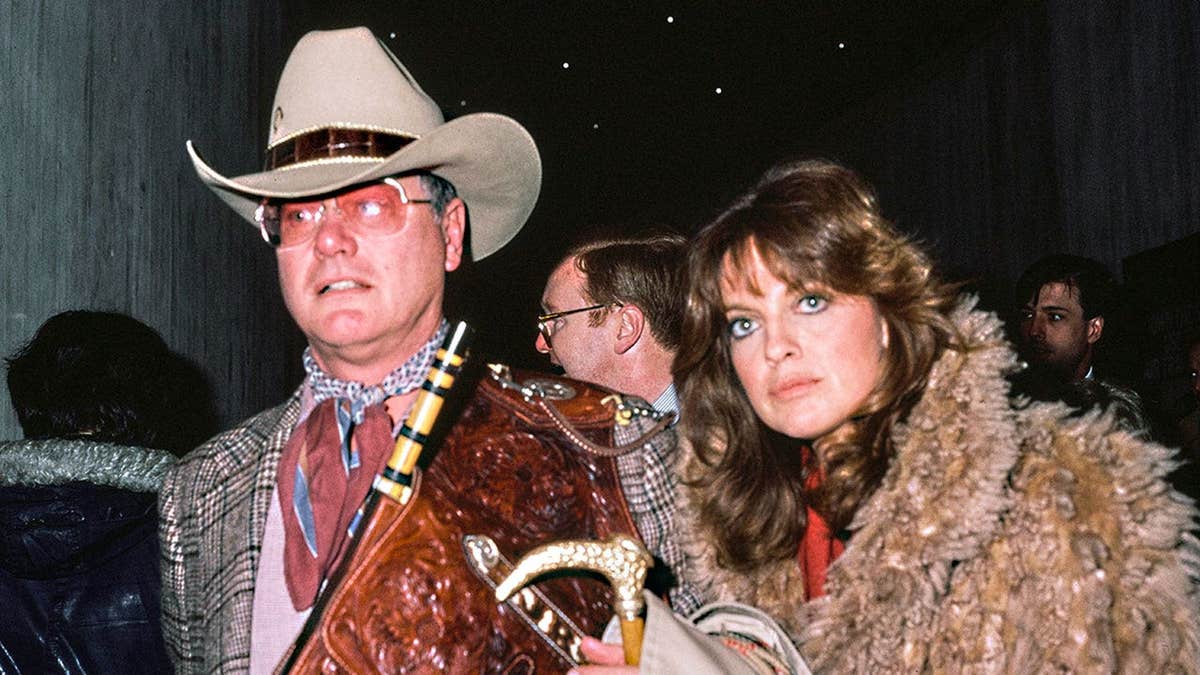 Linda Gray wearing a fur coat walking alongside Larry Hagman in a cowboy hat.