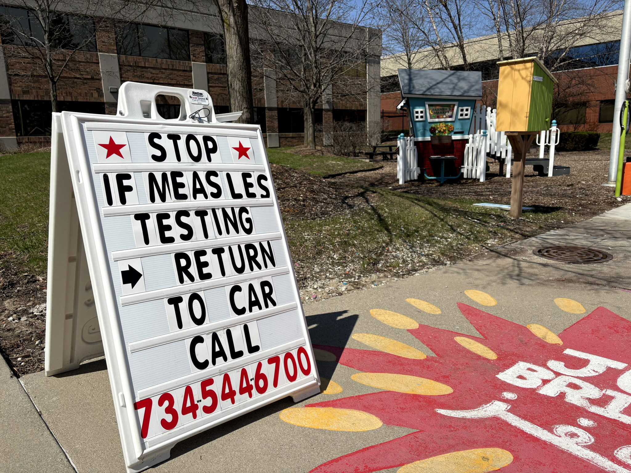 A white sign on a sidewalk says "Stop. If measles testing, return to car. Call 7345446700"