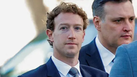 Reuters Mark Zuckerberg wearing a suit, looks towards the camera with a blank expression. He is flanked by staff also in suits.