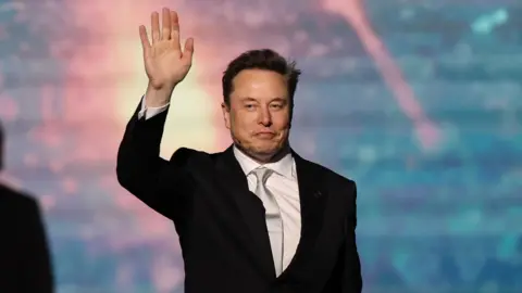 EPA SpaceX chief executive Elon Musk walking onto a stage and waving, wearing a black suit, white collared shirt and shiny off-white neck tie.