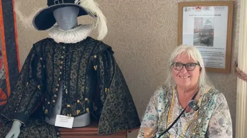 A woman with a multicoloured shirt and black glasses sits next to a black and gold mannequin