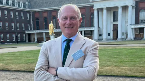 Richard Haldenby Richard Haldenby is standing with his arms crossed wearing a cream jacket and a blue shirt and green tie, arms crossed, in an open courtyard. He has a name badge on his jacket.