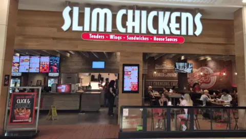 BBC A Slim Chickens store inside a shopping centre with shoppers walking past.