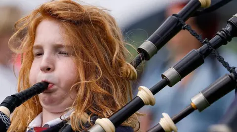 Getty Images Close-up photograph of a teenager playing bagpipes. The piper has long, wavy, red hair and their cheeks are puffed out as they blow into the mouthpiece