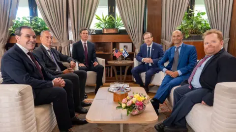 Isaac Herzog/X Isaac Herzog, Jared Kushner and Nickolay Mladenov are among six officials sitting on cream sofas in a wood-panelled room with beige curtains hanging behind them, next to plants placed on window sills. The six men are all wearing suits and ties.