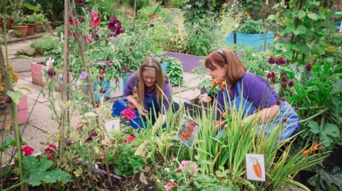 Thrive Two women wearing purple t-shirts and dungarees, smiling and gardening together in a community garden. They are planting vegetables, including carrots and tomatoes, and are surrounded by greenery.