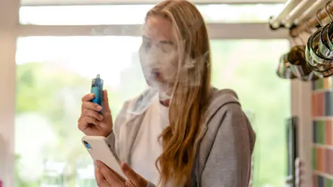 Getty Images A young fair-haired woman wearing a grey hooded sweatshirt and white T shirt is standing in a kitchen with vape and mobile phone in her hands