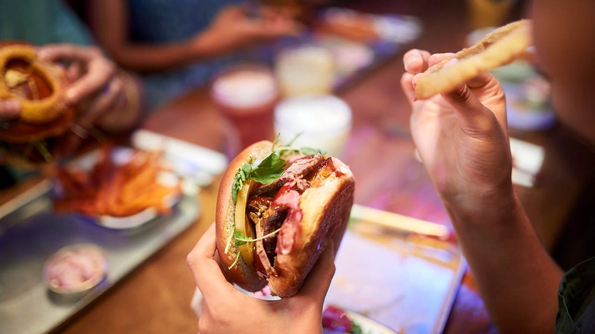 A person holds a burger and takes a bite of a French fry.