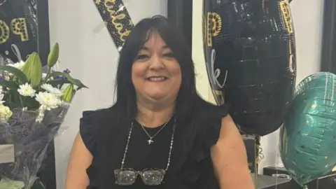 Becca Luttman Louisa Curtis is wearing a sleeveless black top with ruffled detailing and has a pair of glasses hanging from a chain around the neck. Jewellery including a necklace, bracelet, ring and wristwatch, is visible.