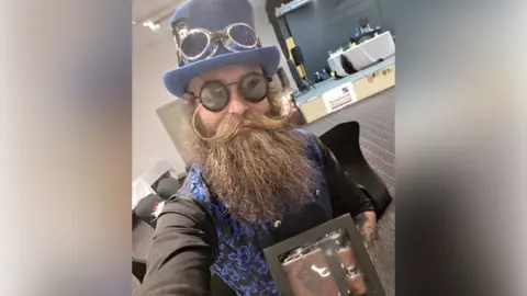 Lewis Aylmer Lewis Aylmer taking a selfie, holding a box of a flask and two shot glasses. He is wearing a black, long-sleeved shirt, a blue and black waistcoat, black glasses and a blue top hat. He has a long, bushy, brown beard and a long, brown moustache.