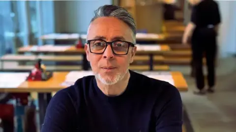 Saviynt Simon Gooch, with quiffed black and grey hair, wears a blue crew neck jumped.