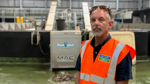 BBC Chris Lofts has curly grey hair and a short grey beard. He wears a dark blue short-sleeved shirt underneath a bright orange hi-vis vest with Severn Trent's logo emblazoned on the front.
