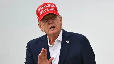Getty Images A close up shot of President Donald Trump speaking to outside with his hand raised. He is wearing a "make America great again" cap