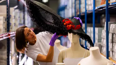 Fashion Museum Bath A woman wearing a white blank t-shirt and purple gloves is angling a large hat that is black with red feathers atop a mannequin