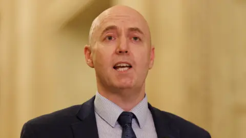 PA Media Jon Burrows is talking. He's wearing a navy suit with a white and blue stripped shirt and a navy tie with a pattern. The background is out of focus.