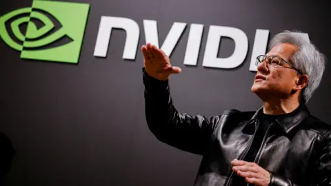 Reuters Nvidia CEO Jensen Huang standing in front of a screen with the company's name and logo, gesturing upward with his hands, wearing a black leather jacket over a black shirt.
