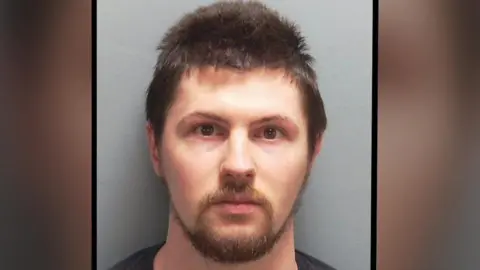 Hampshire Police Custody image of 30-year-old Ethan Orchard. He has short dark hair, dark eyes, light complexion, and a light brown moustache and beard. He is wearing a black t-shirt.