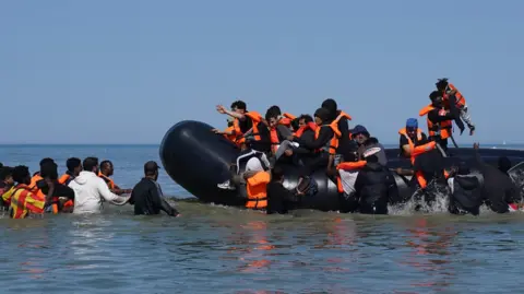 PA Media Migrants climb onto small boat in sea that is around waist-height. They wear orange life jackets