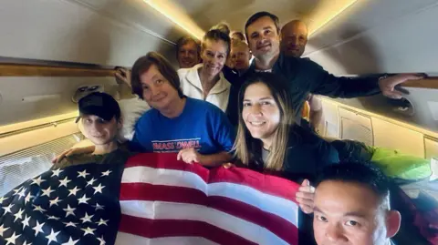 Truthsocial.com/realDonaldTrump Gilman, left, seen with an American flag draped over him on the plane. He is beside several other people, including his mother