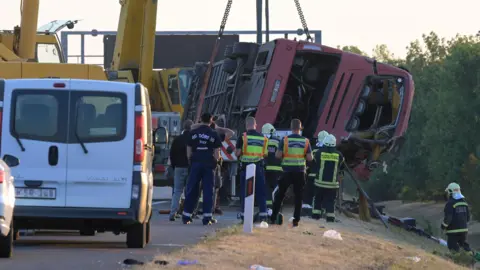 EPA A crane winches an overturned bus up on the side of the road as firefighters look on.
