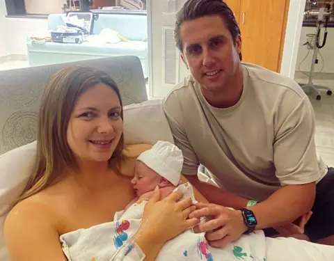 Maisie Savage Newborn baby in the foreground, wearing a white knit hat and wrapped in a white hospital blanket with colourful cartoon prints, held against the chest of a person sitting upright in a hospital bed. An IV cannula and tubing are attached to the person’s forearm. Another person in a light-coloured short-sleeved top sits beside the bed with one hand resting on the blanket and a smartwatch on their wrist. White bedding, pillows and a hospital room with cabinets, a sofa, medical equipment and windows are visible in the background.