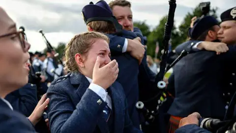 Getty Images An emotional female piper, wearing a traditional outfit and with her hair in a French plait, is standing with one hand covering her mouth and part of her face. She is surrounded by other pipers who are hugging each other