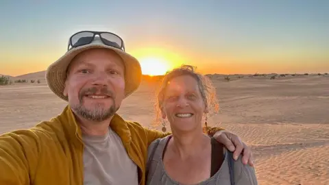 Supplied by Nick Smith A selfie taken by Nick Smith showing him wearing a yellow jacket and a beige undershirt, and a beige hat with sunglasses sitting on top of his head, his arm around his wife, who is smiling and has an updo and is wearing dangling earrings. Behind them is the sun setting over the Moroccan desert.