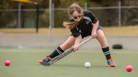 Supplied A girl wearing black sports shorts and t-shirt leans forward to hit a white ball with a hockey stick
