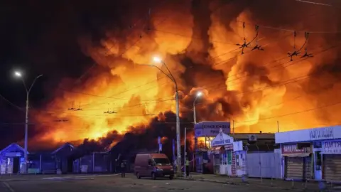 Reuters Large fire engulfs a market area at night, sending towering flames and thick smoke into the sky above nearby buildings, streets, and overhead power lines.