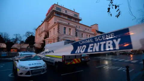 CAROLINE QUINN/GETTY IMAGES A garda (Irish police) cordon outside the Regency Hotel in Dublin in 2016. A white and blue liveried garda van and car are parked outside a four-storey hotel at dusk on a wet evening. A white a blue cordon tape stretches across an entrance to the hotel car park.