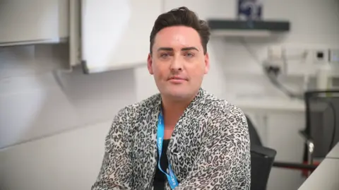 Jamie Niblock/BBC Jamie is sitting in a chair in an office he is wearing a leopard print shirt and black top underneath.