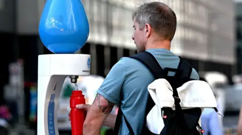 Getty Images Back view of a man wearing a blue t-shirt and black backpack filling up a red water bottle at a temporary drinking fountain