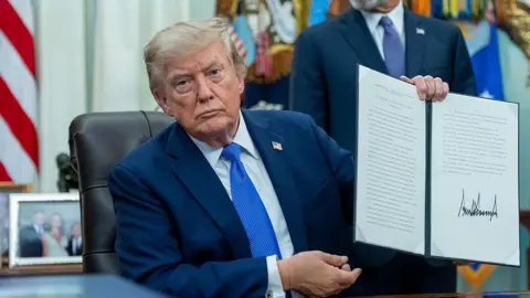 Bloomberg via Getty Images Trump, wearing a blue suit and sitting at desk in the Oval Office, holding up a signed executive order