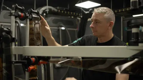 BBC A man with silver hair adjusts a ruler over which film is looped