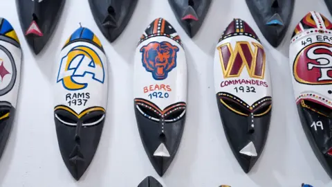 A row of traditional Ghanaian masks with the logos and names of football teams - the Rams, the Bears and the Commanders, painted on