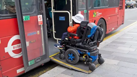 Alamy A person in a motorised wheelchair wearing an orange jacket and a white hat boards a bus in London using a wheelchair ramp in October 2025