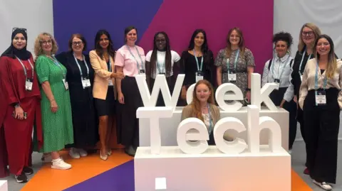 TechSY Eleven smiling colourfully dressed women, wearing lanyards are posed behind large white 3D 'Tech Week' signage with another woman seated between the words 'Tech' in the foreground, and 'Week', in front of the row of smiling women.