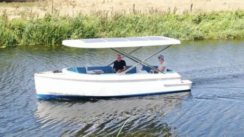 Torkmar Tim Knox and his wife Sue travelling on the boat. It is a small white vessel with solar paneling on its roof.
