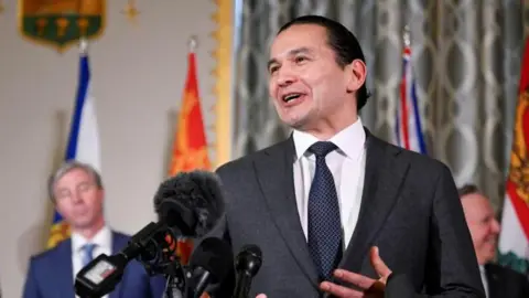 Reuters An image showing Wab Kinew speaking in front of several microphones at a press conference with a few of Canada's other premiers behind him. He has black hair that is tied up in a bun, and is wearing a dark charcoal grey suit and tie.