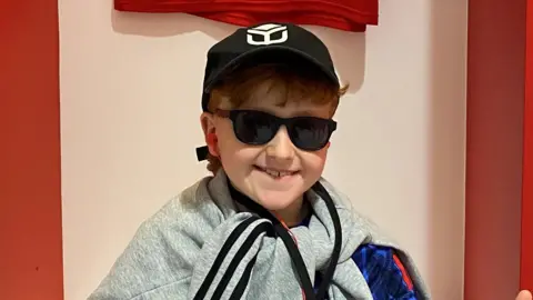 Contributed A young boy sits in a football changing room smiling at the camera. He has both thumbs raised up. He wears a black cap and sunglasses and has a grey jumper tied around his neck.