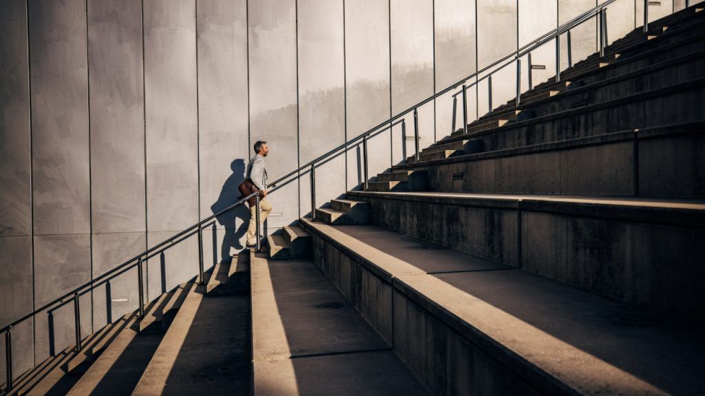 A middle-aged adult dressed in business attire climbs a flight of stairs during evening sun