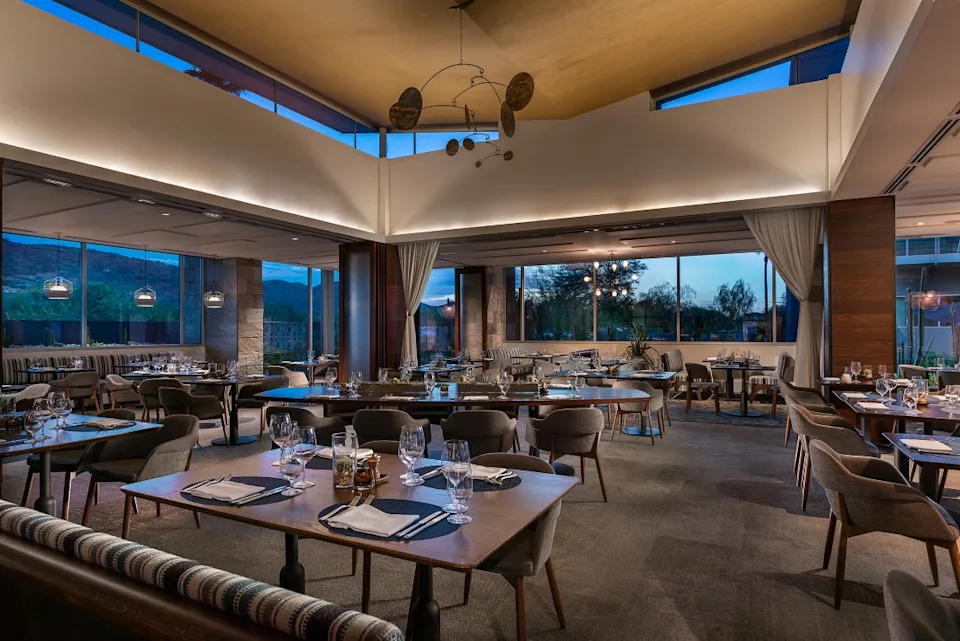 A dining room at dusk. All the tables are set with silverware and glasses