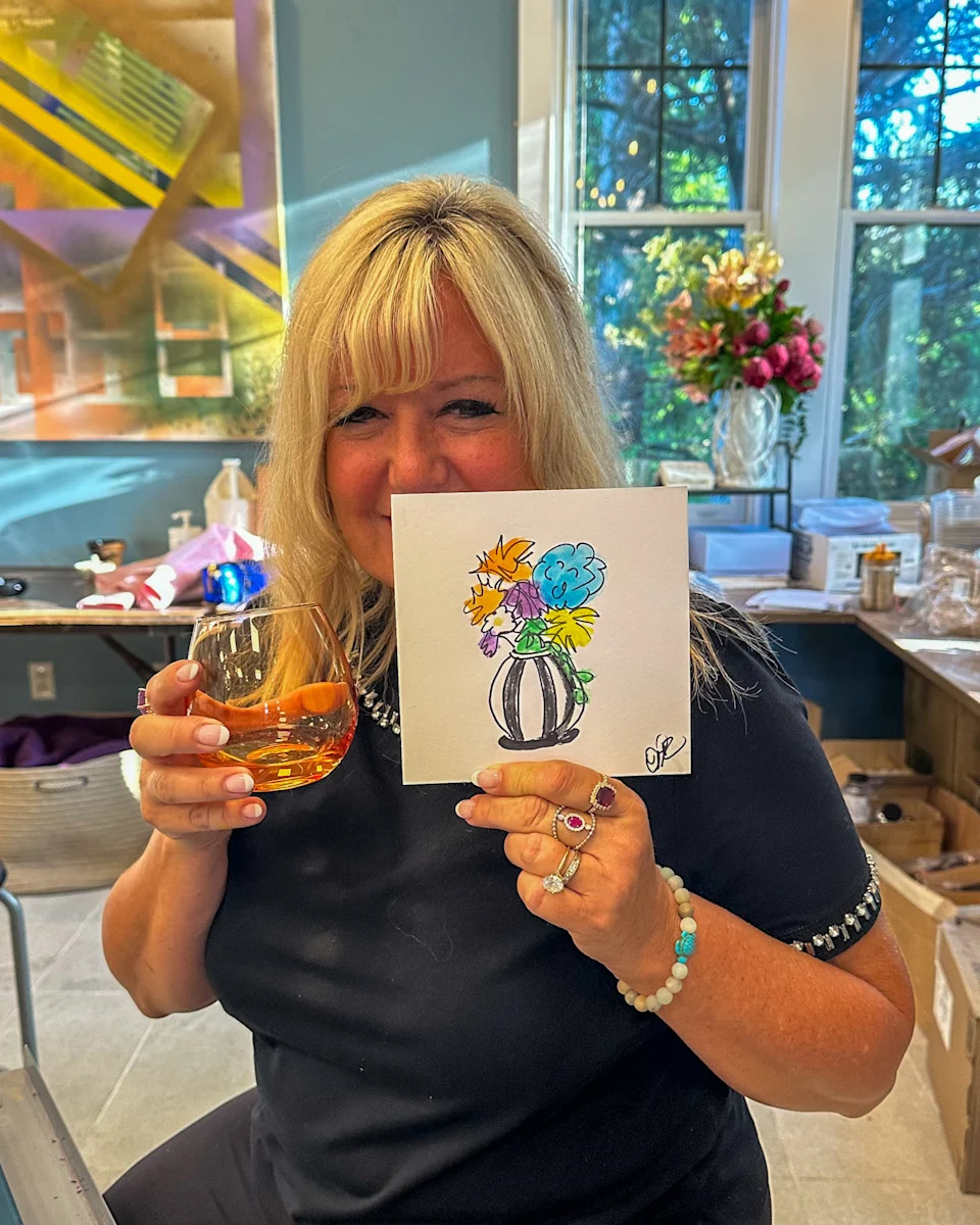 A woman holds a glass of wine and a painted photo of a vase with flowers